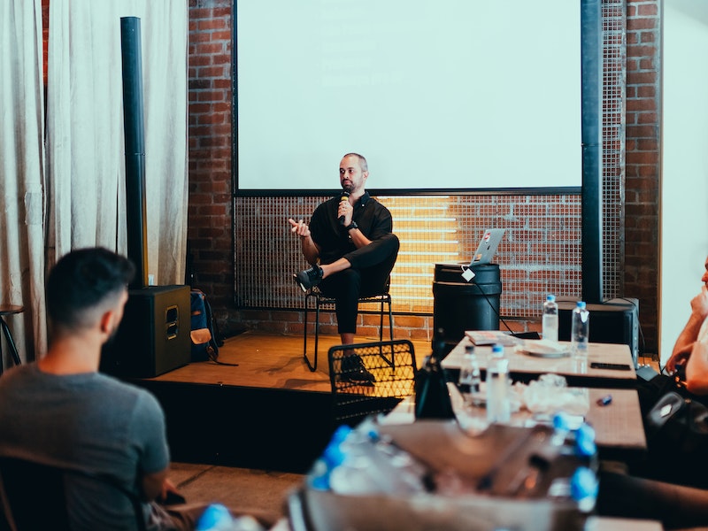 Person presenting in brick-walled room with projector screen and audience.