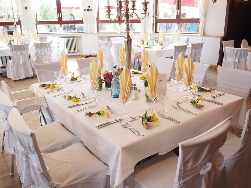 Table setting in restaurant with white tablecloth, yellow runner, white plates, silverware, wine glasses, and yellow napkins.