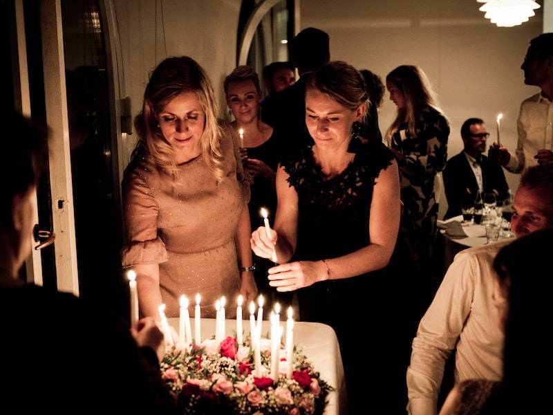 Group of people celebrating around a large cake with red and pink roses and white candles in a dimly lit room.