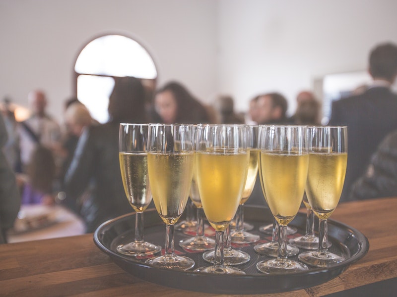 Tray of champagne glasses on wooden table in crowded room with arched window.