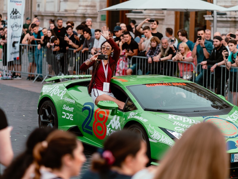 Green race car with number 28 and Kappa logo driving on cobblestone street with crowd.