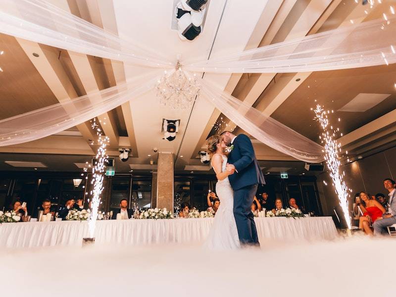 Romantic couple dancing and kissing in ballroom with chandelier and sparklers, white dress and navy suit.