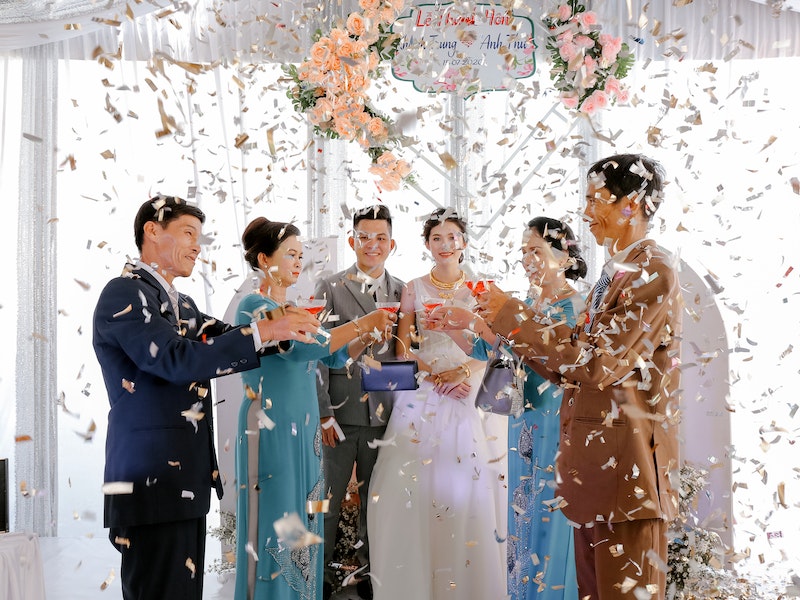 Group of people celebrating at Le Nguyen Nhu Y wedding day under white flower arch with champagne glasses and confetti falling.