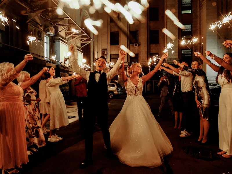 Newlywed couple dancing in street at night surrounded by people with sparklers, warm and celebratory mood.