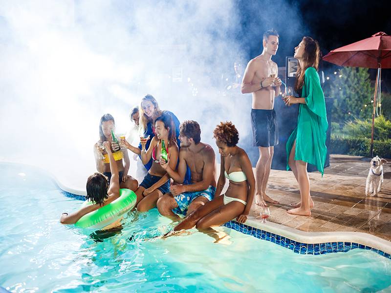 Group of people socializing in pool at night with drinks and dog on stone deck under red umbrella.